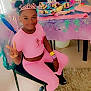 child, girl, birthday_party, tiara, pink_clothing, peace_sign, cake, table, chair, wristbands, smile, celebration, indoor, carpet, party_decor, food, happy, person, festive, hair_buns