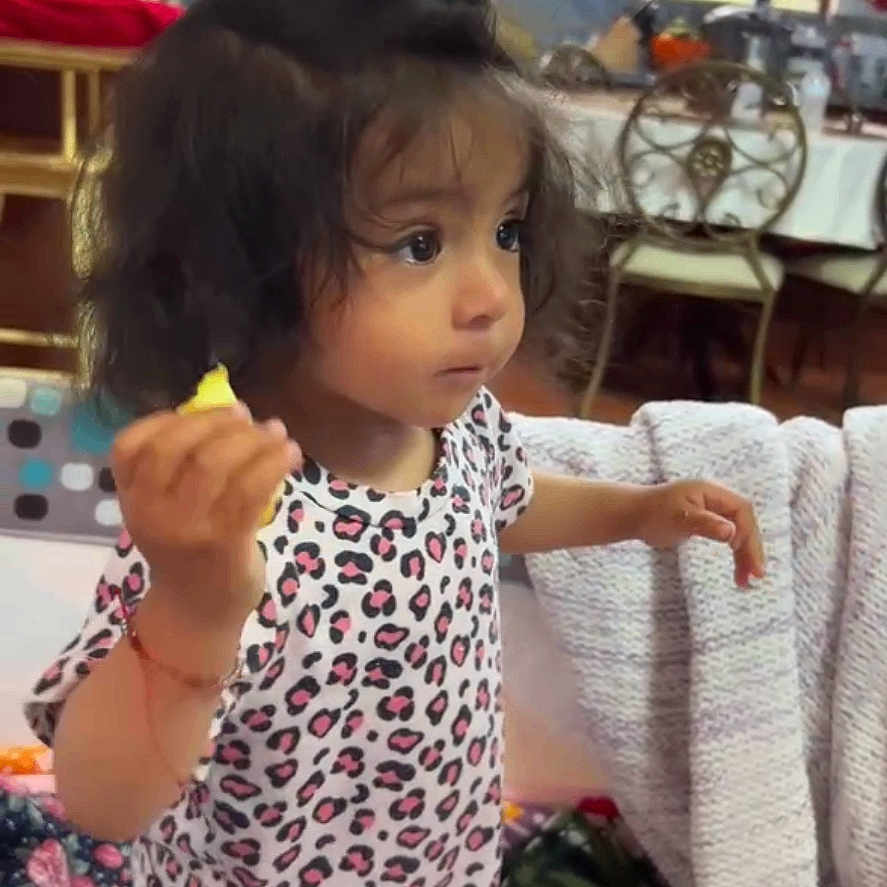 Danna joined the competition — help win amazing prizes! blanket, casual, chair, child, clothing, curly_hair, curtain, cushion, face, furniture, holding_object, home, indoor, patterned_shirt, person, phone_screen, pink_pants, toddler, video_call, window