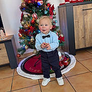 Layden a rejoint le concours — aidez-le/la à gagner de superbes lots ! toddler, child, christmas_tree, holiday_decorations, ornaments, bowtie, blue_shirt, white_shoes, tile_floor, indoor, cabinet, festive, train_set, santa_figurine, red_bow, pine_tree, smiling, standing, toy, holiday