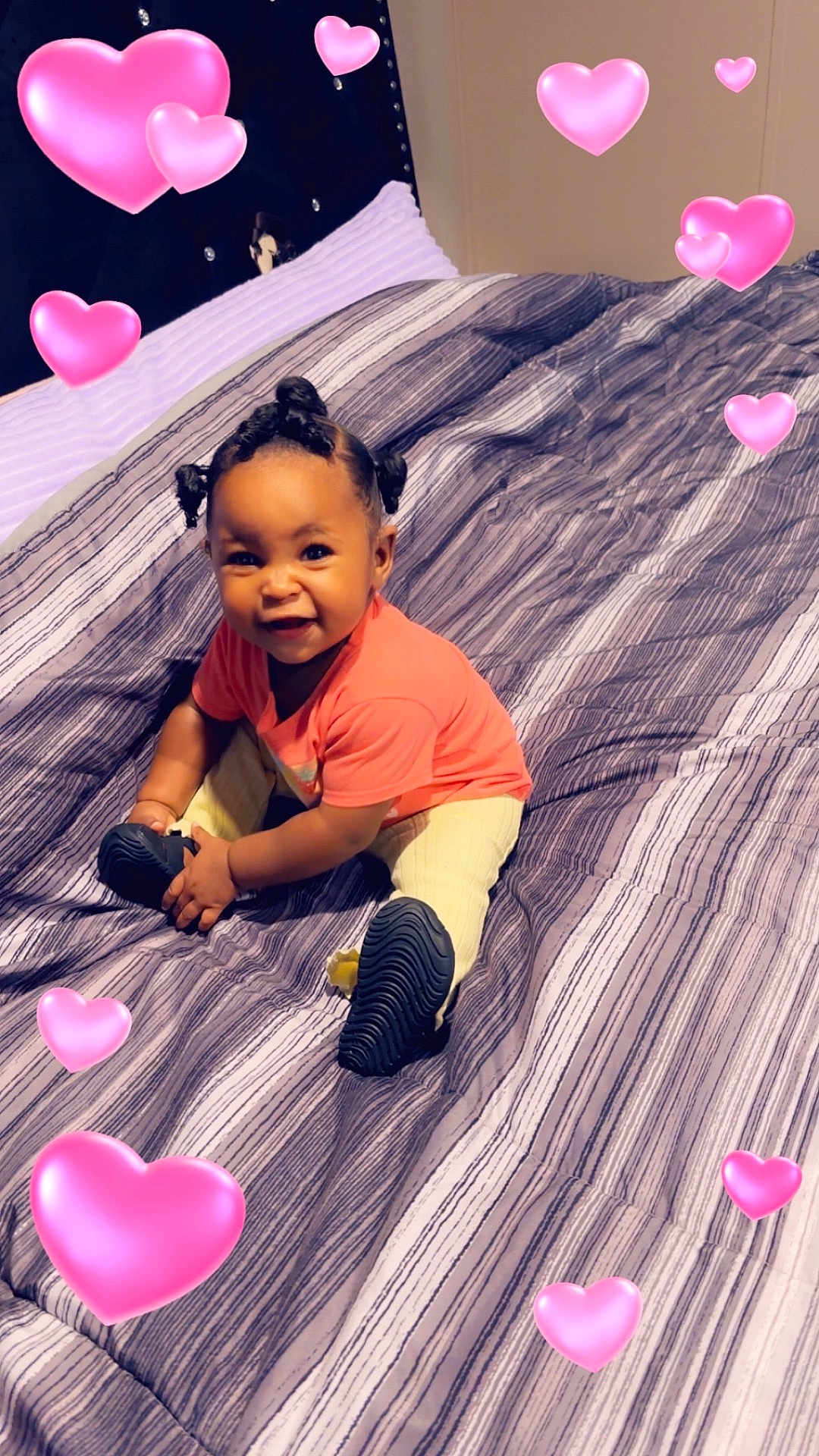 Reign is registered to the contest to win money with this photo: baby, child, smiling, sitting, bed, striped_bedspread, indoor, hair_buns, pink_shirt, yellow_pants, shoes, happy, cute, portrait, heart_emojis, cozy, room, playful, infant, young_child