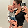baby, woman, mirror_selfie, kissing, holding, indoor, casual_clothing, black_top, denim_shorts, floral_pattern, hair_bun, smartphone, nail_polish, reflection, affection, parent_and_child, portrait, skin, hand, face