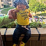 baby, child, hat, yellow_shirt, text_on_clothing, hand, flowers, balcony, plants, sunlight, outdoor, building, apartment, greenery, jeans, yellow_shoes, person, portrait, cute, smiling