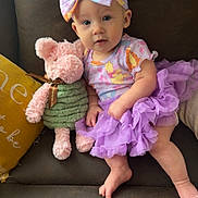 Ivyanna is registered to the contest to win money with this photo: baby, child, tutu, headband, stuffed_animal, plush_toy, couch, pillow, purple, pink, yellow, clothing, sitting, indoor, cute, infant, person, soft_toy, leather, smiling