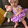 baby, child, tutu, headband, stuffed_animal, plush_toy, couch, pillow, purple, pink, yellow, clothing, sitting, indoor, cute, infant, person, soft_toy, leather, smiling