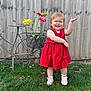 toddler, child, red_dress, grass, flower_pot, flowers, metal_bicycle, fence, outdoor, smiling, playful, greenery, yard, plant, person, footwear, socks, happy, garden, summer