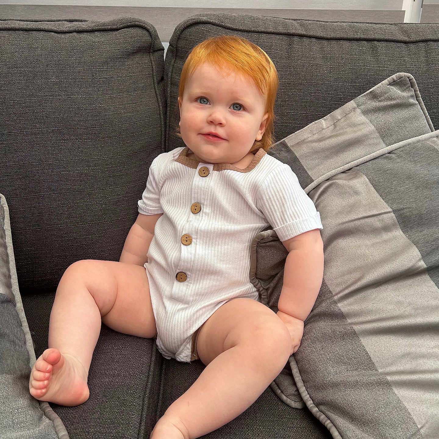 Amelie is registered to the contest to win money with this photo: baby, barefoot, blue_eyes, buttoned_clothing, child, comfortable, couch, curious, cushions, home, indoor, light, onesie, plush, portrait, red_hair, sitting, smiling, soft_texture, toddler