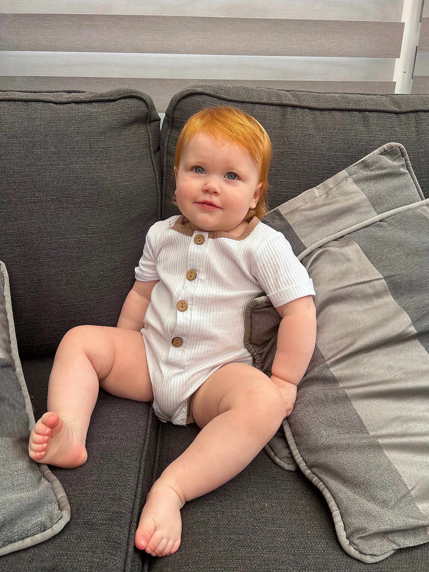 Amelie is registered to the contest to win money with this photo: toddler, child, red_hair, blue_eyes, couch, cushions, indoor, onesie, buttoned_clothing, barefoot, plush, comfortable, home, portrait, sitting, smiling, curious, baby, soft_texture, light
