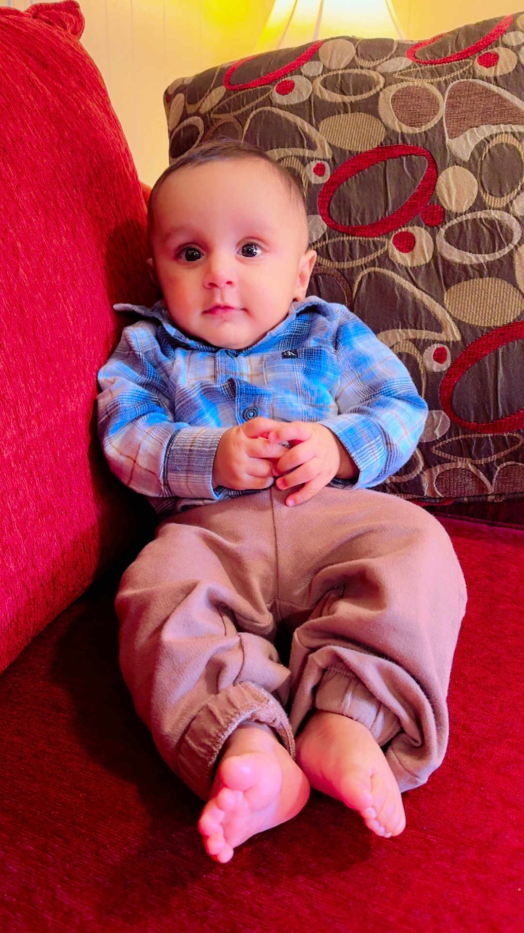 Priyansh Minhas joined the competition — help win amazing prizes! baby, child, infant, couch, plaid_shirt, patterned_cushion, red_couch, beige_pants, barefoot, indoor, portrait, sitting, cute, hands, feet, young_child, comfortable, curious, furniture, home
