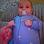 baby, pacifier, blue_onesie, bee_pattern, hand, finger, cushion, blanket, floor, carpet, indoor, person, infant, child, smiling, closeup, cozy, cute, sleepwear, relaxed