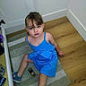 child, girl, blue_dress, sitting, rug, floor, perfume_bottle, indoor, corner, wood_floor, curious_expression, person, young_child, casual, footwear, hair, face, eyebrows, lip_gloss, hand