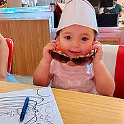 Alessi joined the competition — help win amazing prizes! toddler, child, paper_hat, sunglasses, coloring_sheet, crayons, table, diner, restaurant, wooden_panel, person, seat, red_chair, white_shirt, smiling, cute, indoor, face, hands, toy