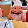 toddler, child, paper_hat, sunglasses, coloring_sheet, crayons, table, diner, restaurant, wooden_panel, person, seat, red_chair, white_shirt, smiling, cute, indoor, face, hands, toy