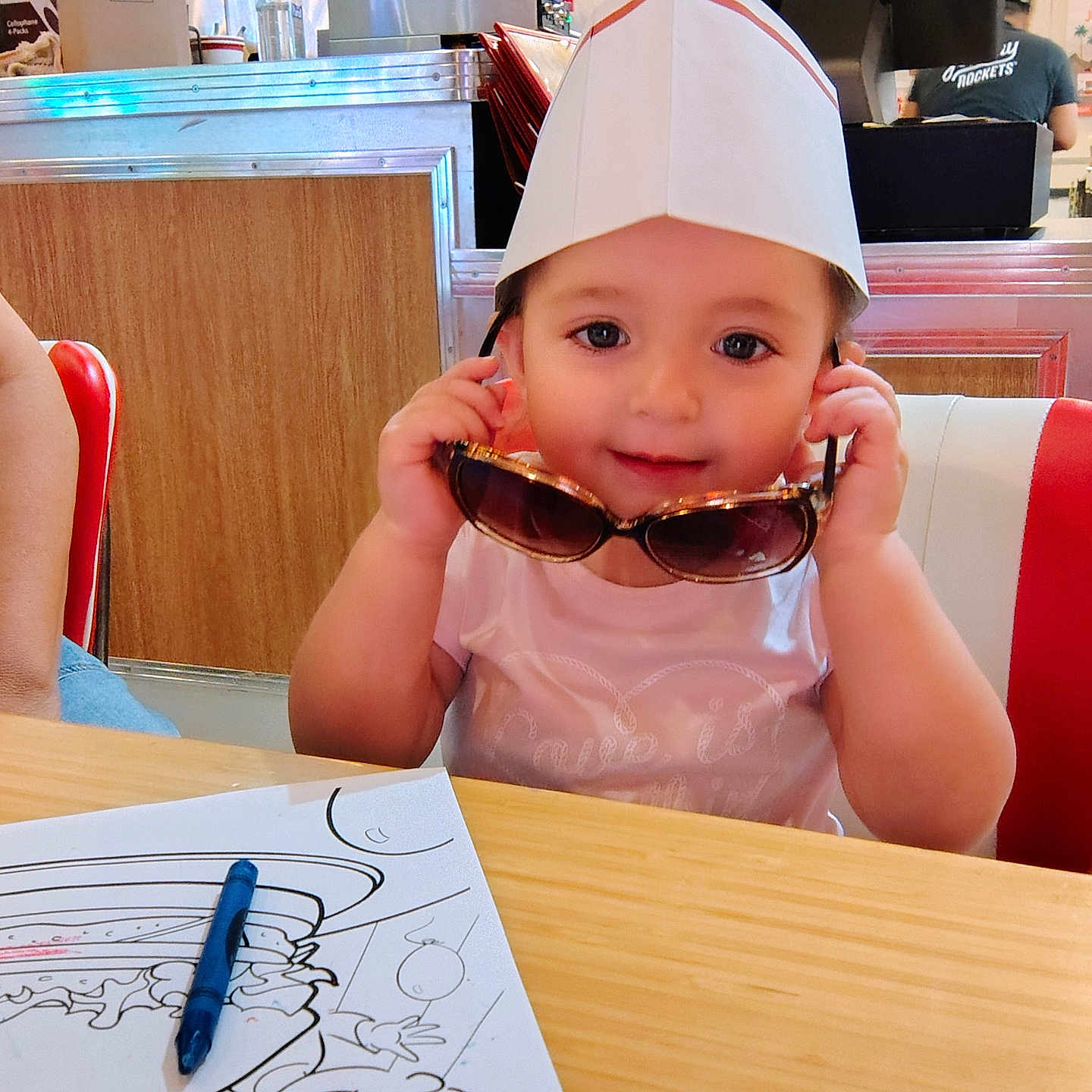 Alessi joined the competition — help win amazing prizes! toddler, child, paper_hat, sunglasses, coloring_sheet, crayons, table, diner, restaurant, wooden_panel, person, seat, red_chair, white_shirt, smiling, cute, indoor, face, hands, toy