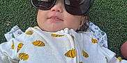 Rubyrae is registered to the contest to win money with this photo: baby, child, sunglasses, grass, outdoor, onesie, bee_pattern, cute, infant, face, smile, bubble, clothing, relaxed, lying_down, toddler, nature, sunlight, head, portrait