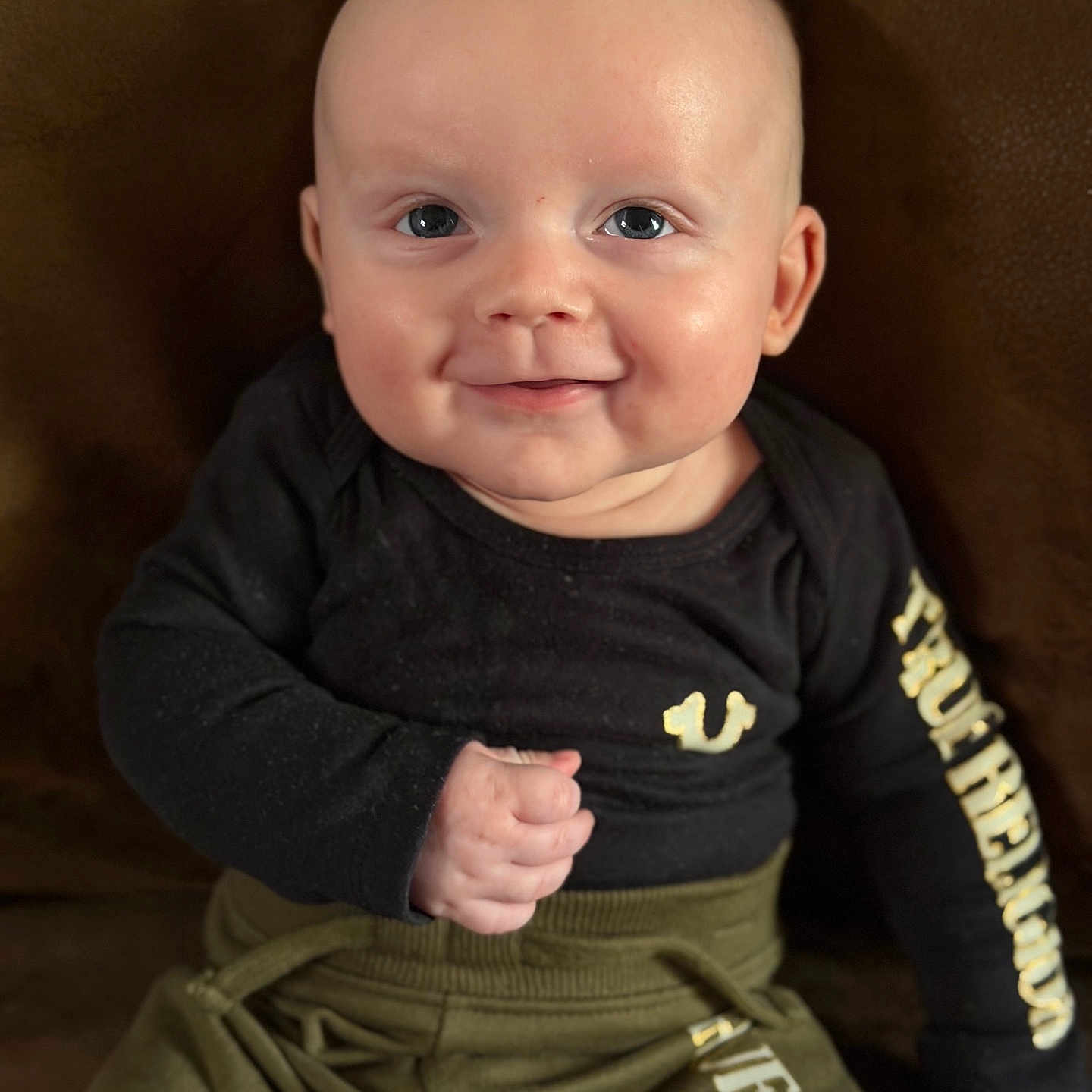 Maverick joined the competition — help win amazing prizes! baby, smiling, face, child, clothing, black_shirt, olive_pants, sitting, fist, happy, portrait, indoor, infant, cute, young_child, person, front_view, bright_eyes, human, casual_wear
