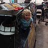 child, curly_hair, blue_shirt, race_car, garage, tires, chair, tools, indoor, vehicle, playful, person, window, cluttered, mechanical, floor, sign, poster, helmet, racing