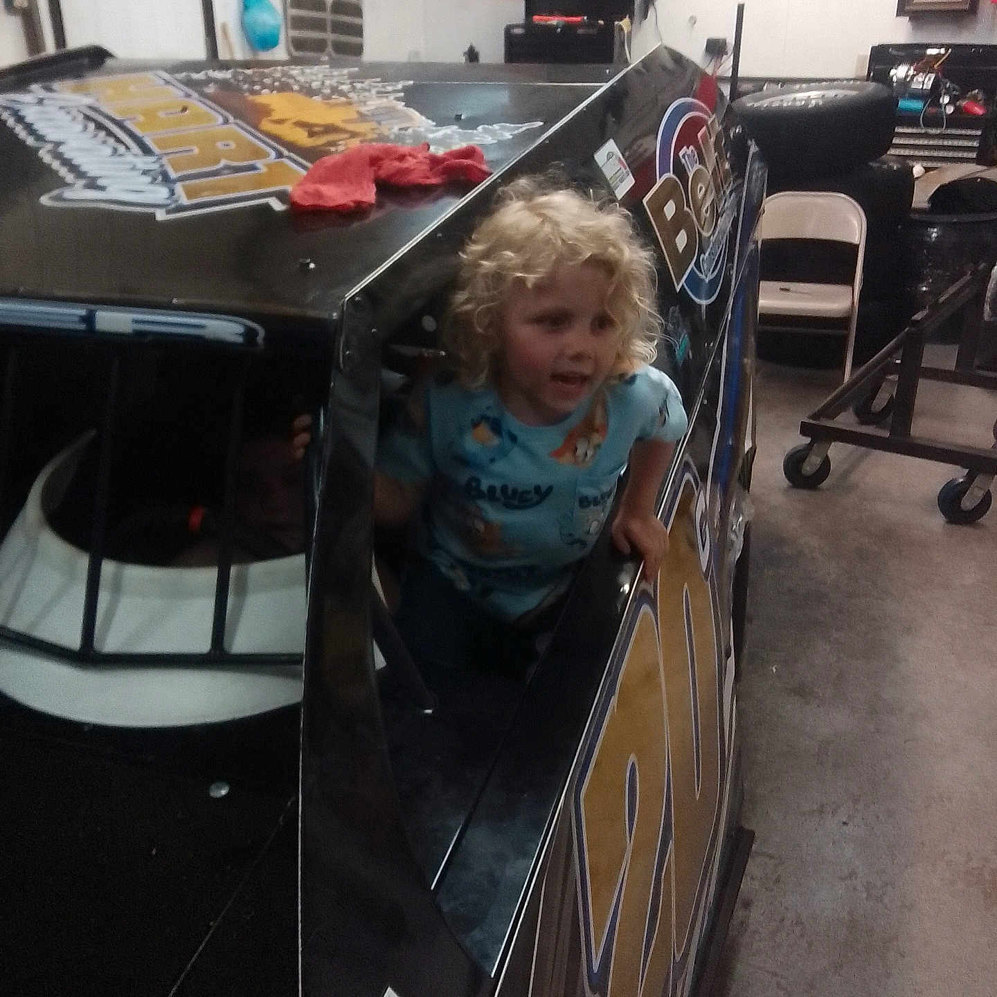 Raiden joined the competition — help win amazing prizes! child, curly_hair, blue_shirt, race_car, garage, tires, chair, tools, indoor, vehicle, playful, person, window, cluttered, mechanical, floor, sign, poster, helmet, racing
