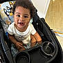 toddler, child, smiling, curly_hair, stroller, indoor, baby_clothes, seat, happy, person, cute, playful, cushion, toy_pattern, black_hair, skin, infant, young_child, sitting, bright