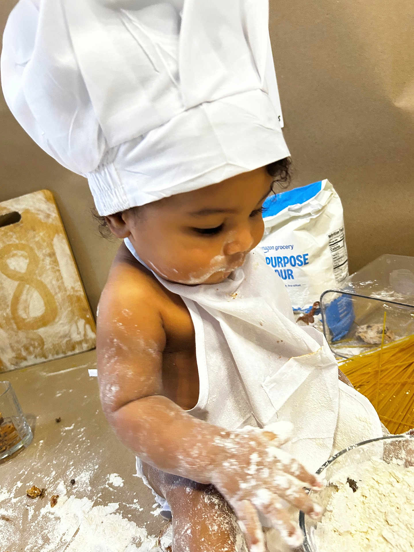 Aiden joined the competition — help win amazing prizes! baby, child, chef_hat, apron, flour, baking, dough, kitchen, messy, cooking, food_preparation, table, spoon, ingredient, closeup, person, indoors, cute, fun, playful