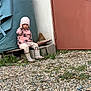 beanie, boots, casual_clothing, child, concrete, curious, daylight, door, footwear, gravel, leopard_print, nature, outdoor, pink, plant, rust, sitting, small, tarp, wall
