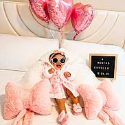 Fiorella is registered to the contest to win money with this photo: baby, child, pink, robe, sunglasses, bed, pillows, balloons, heart_shaped, plush, slippers, sign, smiling, cute, indoor, cozy, celebration, headband, person, happy