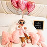 baby, child, pink, robe, sunglasses, bed, pillows, balloons, heart_shaped, plush, slippers, sign, smiling, cute, indoor, cozy, celebration, headband, person, happy