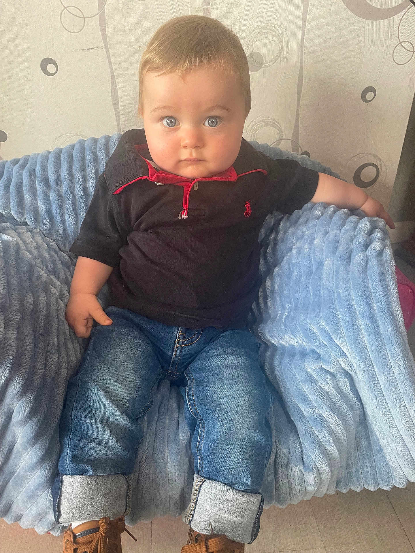 Mylo a rejoint le concours — aidez-le/la à gagner de superbes lots ! baby, child, blue_eyes, black_polo_shirt, jeans, brown_shoes, plush_chair, indoor, curious, serious_expression, cute, infant, casual_clothing, cozy, seated, portrait, human, young_child, floor, wallpaper