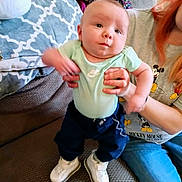 Braun is registered to the contest to win money with this photo: adult, baby, child, clothing, couch, footwear, hand, holding, indoor, infant, jeans, living_room, mickey_mouse, motion_blur, pants, pattern, person, pillow, shirt, sneakers