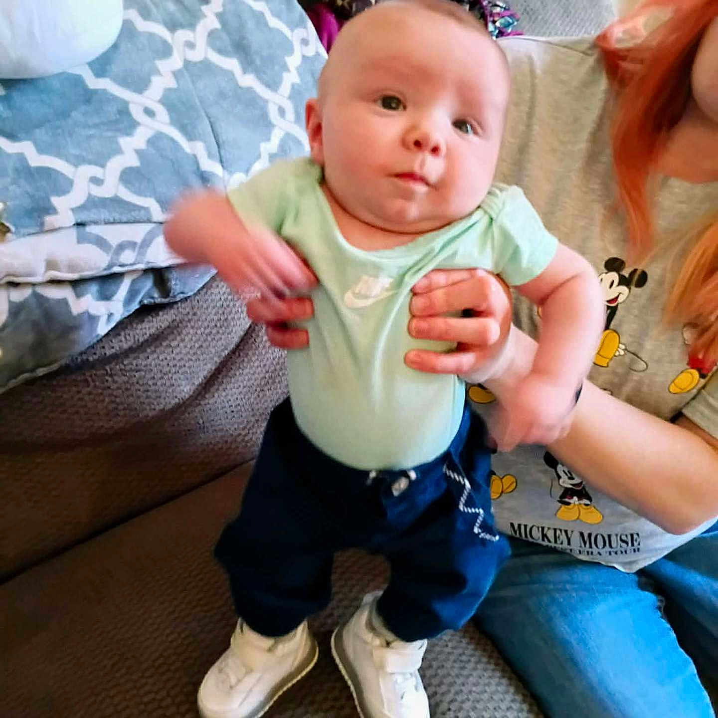 Braun is registered to the contest to win money with this photo: adult, baby, child, clothing, couch, footwear, hand, holding, indoor, infant, jeans, living_room, mickey_mouse, motion_blur, pants, pattern, person, pillow, shirt, sneakers