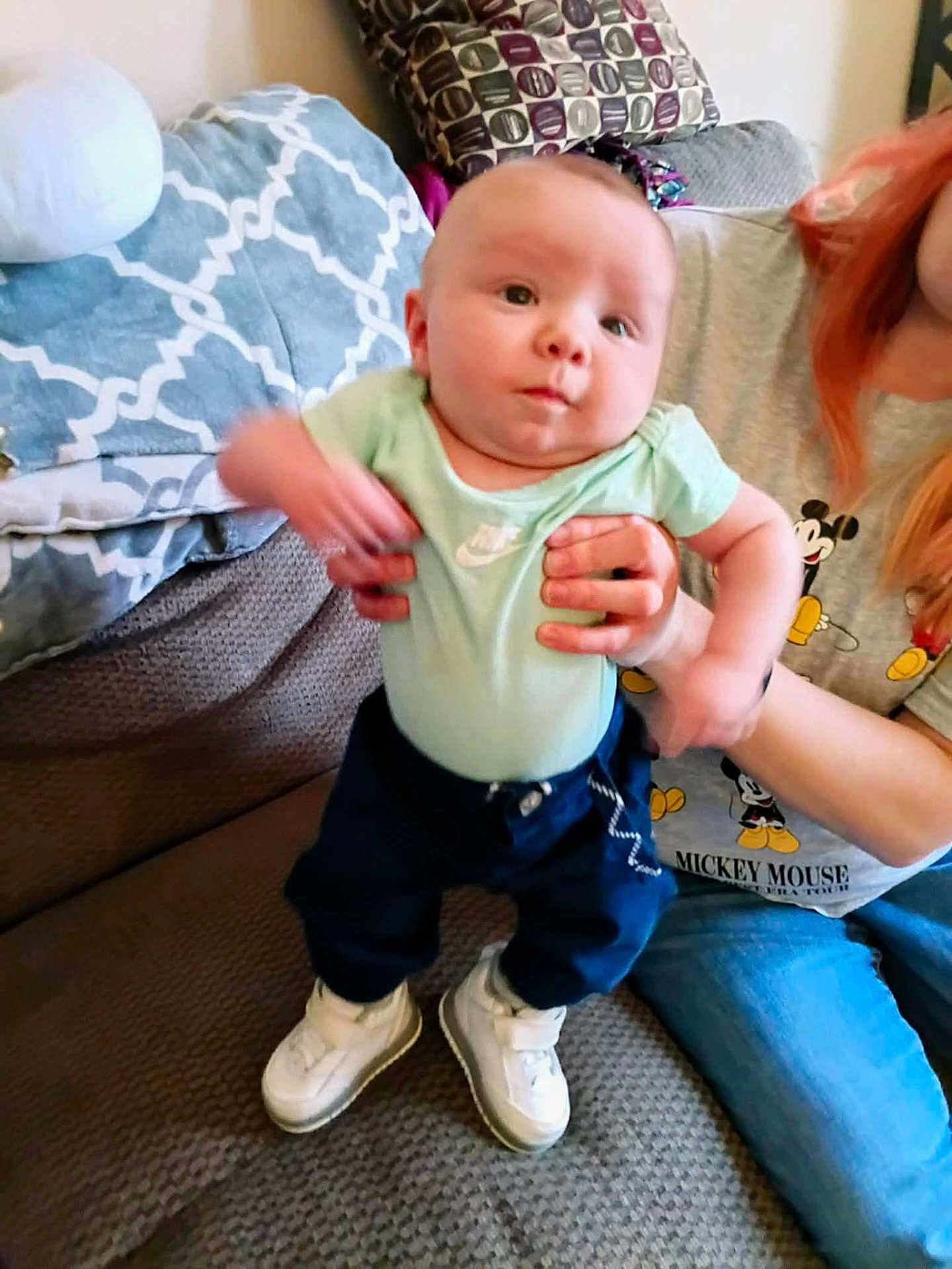 Braun is registered to the contest to win money with this photo: adult, baby, child, clothing, couch, footwear, hand, holding, indoor, infant, jeans, living_room, mickey_mouse, motion_blur, pants, pattern, person, pillow, shirt, sneakers