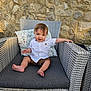 Oscar is registered to the contest to win money with this photo: baby, child, smiling, white_shirt, brown_shorts, wicker_chair, outdoor, stone_wall, cushion, floral_pattern, barefoot, happy, person, sitting, cute, portrait, infant, sunlight, relaxed, casual