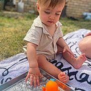 Oscar joined the competition — help win amazing prizes! toddler, child, water, outdoor, toy, orange_octopus, blue_cup, grass, towel, hand, foot, short_sleeve_shirt, playing, summer, sunlight, brick_wall, curious, sitting, baby, water_play