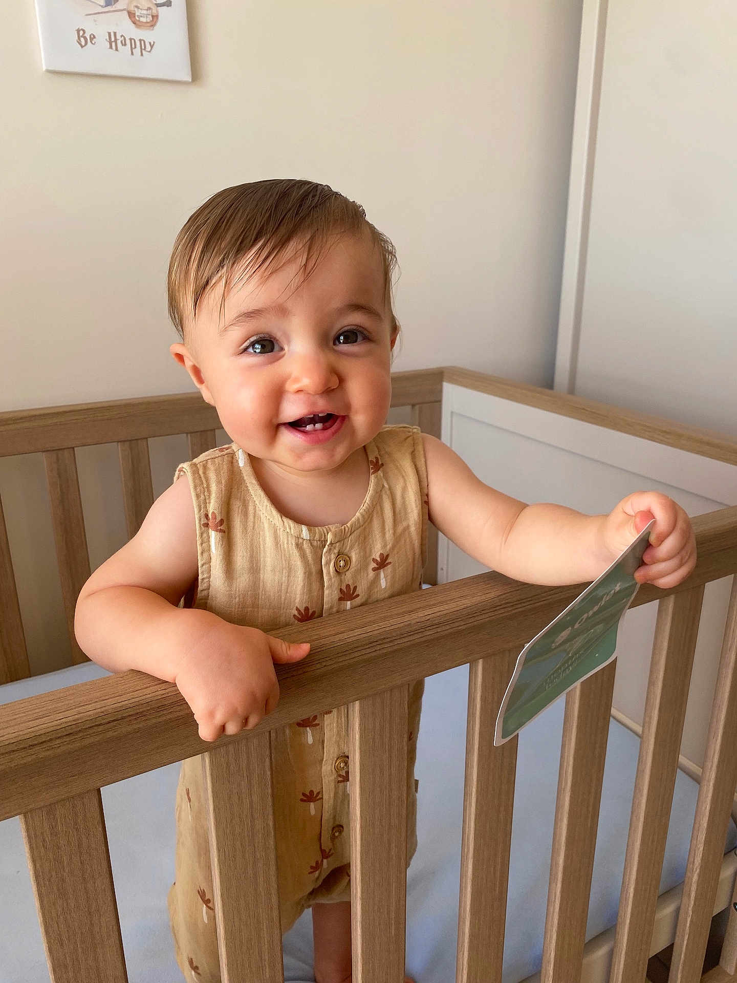 Oscar is registered to the contest to win money with this photo: toddler, child, crib, wooden_crib, smiling, happy, indoor, card, baby_clothes, nursery, person, face, hand, hair, buttoned_outfit, pale_wall, wood, infant, cute, holding_object