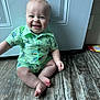 Dawson is registered to the contest to win money with this photo: baby, blue_eyes, bright, child, cute, door, flooring, green_clothing, happy, home, indoor, infant, patterned_clothing, person, shirt, shorts, sitting, smiling, wooden_floor, young_child