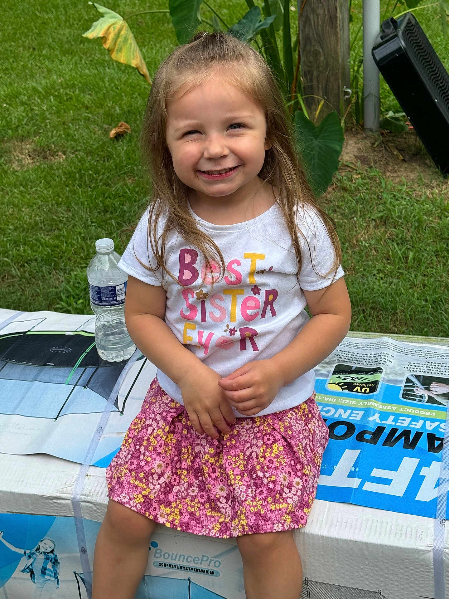 Gracelyn is registered to the contest to win money with this photo: child, girl, smiling, sitting, outdoor, grass, plant, leaf, water_bottle, box, clothing, skirt, tshirt, happy, person, portrait, daylight, nature, casual, cute