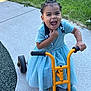 child, girl, tricycle, blue_dress, braids, beads, outdoor, sidewalk, grass, happy, playful, toy, sneakers, smiling, young_child, summer, daylight, expression, joy, recreation