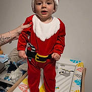 Arthur a rejoint le concours — aidez-le/la à gagner de superbes lots ! baby, baby_changing_pad, child, christmas_costume, cute, footwear, furniture, giraffe_print, hand, home, indoor, person, red_clothing, santa_hat, smiling, standing, support, tattoo, toddler, white_wall