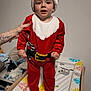 baby, baby_changing_pad, child, christmas_costume, cute, footwear, furniture, giraffe_print, hand, home, indoor, person, red_clothing, santa_hat, smiling, standing, support, tattoo, toddler, white_wall