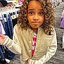 apparel, casual_wear, child, clothing_store, curious_expression, curly_hair, floor, hand_gesture, indoor, jacket, jeans, long_sleeves, person, portrait, rack, retail, shopping, standing, store_aisle, young