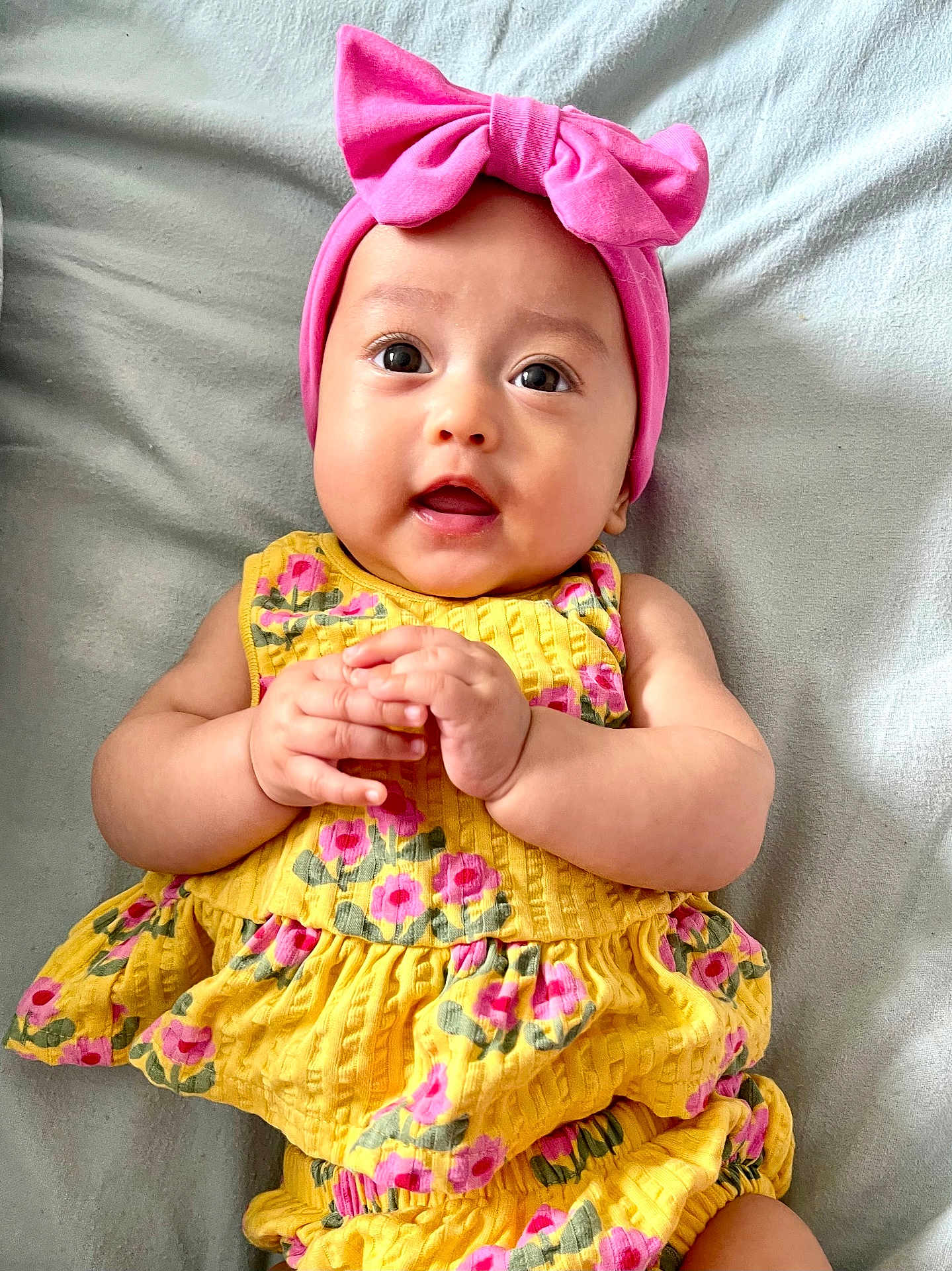 Matilda is registered to the contest to win money with this photo: baby, infant, child, pink_bow, headband, yellow_dress, floral_pattern, hands, clasped_hands, lying_down, gray_blanket, cute, adorable, portrait, happy, smiling, skin, eyes, closeup, soft