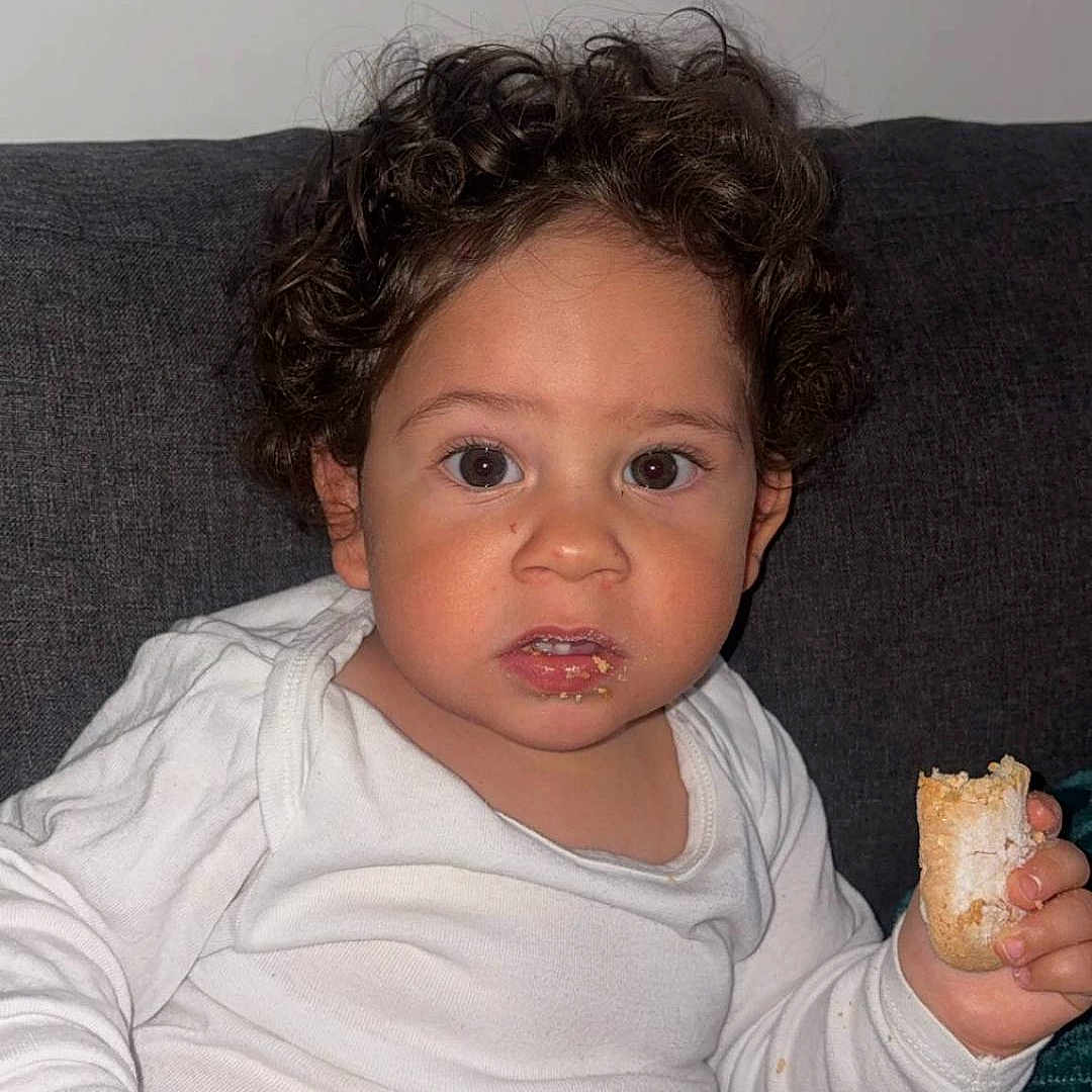 Lévyn participe au concours pour gagner de l'argent avec cette photo : black_pants, casual_clothing, child, couch, crumbs, curly_hair, expression, face, food, hand, indoor, looking_at_camera, pastry, portrait, powdered_sugar, sitting, snack, toddler, white_shirt, young_child