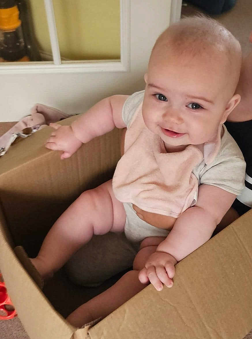 Scarlett joined the competition — help win amazing prizes! baby, cardboard_box, indoor, smiling, bib, cute, child, person, sitting, chubby, happy, skin, face, hand, arm, leg, clothing, door, floor, household