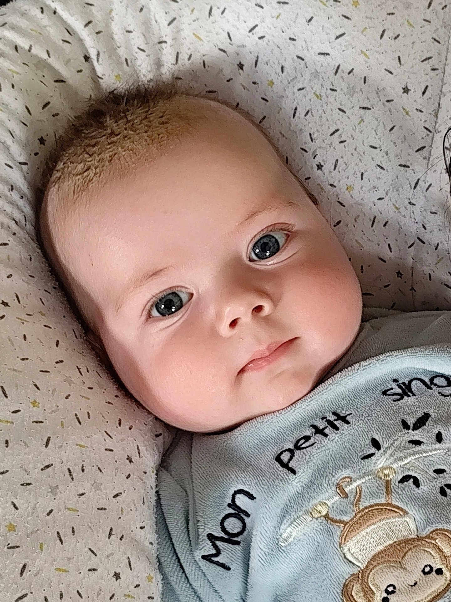 Kézio participe au concours pour gagner de l'argent avec cette photo : baby, infant, face, closeup, blue_eyes, cheeks, clothing, pillow, pattern, cute, soft_lighting, monkey_design, text_on_clothing, portrait, child, young, indoors, laying_down, expression, adorable