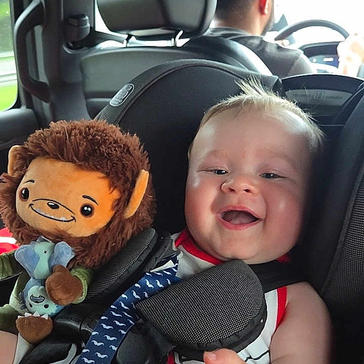 Max is registered to the contest to win money with this photo: baby, child, infant, smiling, happy, car, car_seat, passenger, driver, adult, plush_toy, monkey_toy, toy, vehicle, indoors, travel, portrait, seated, harness, family
