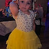child, girl, toddler, birthday, birthday_party, yellow_dress, tutu, skirt, portrait, standing, smiling, face, bow, earrings, indoor, home, decorations, gifts, television, colorful