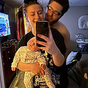 Landen is registered to the contest to win money with this photo: family, baby, woman, man, selfie, mirror, sleeping, smile, indoor, bedroom, television, clothing, holding, casual, hair_bun, glasses, furniture, flooring, device, affection