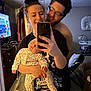 family, baby, woman, man, selfie, mirror, sleeping, smile, indoor, bedroom, television, clothing, holding, casual, hair_bun, glasses, furniture, flooring, device, affection