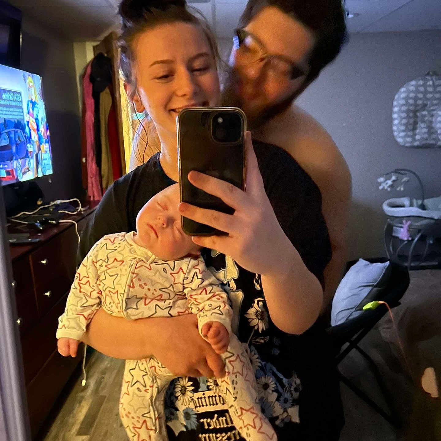 Landen is registered to the contest to win money with this photo: affection, baby, bedroom, casual, clothing, device, family, flooring, furniture, glasses, hair_bun, holding, indoor, man, mirror, selfie, sleeping, smile, television, woman