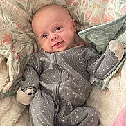 Aviana is registered to the contest to win money with this photo: baby, infant, smile, onesie, grey_clothing, polka_dot, blanket, floral_pattern, soft_texture, cute, happy, lying_down, child, face, head, small_hands, cozy, comfort, indoor, portrait