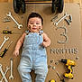 baby, infant, denim_overalls, tools, wrenches, screwdrivers, drill, nuts, bolts, cardboard, face_paint, baby_milestone, measurement_tape, wood_floor, baby_pose, workshop_theme, cute, smiling, indoor, portrait